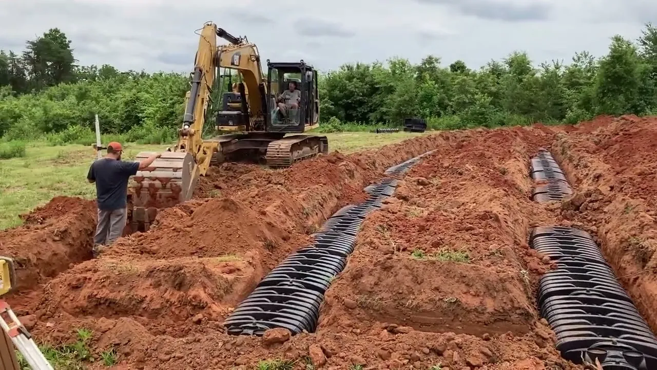 A chamber drain field going in: rows of black plastic septic chambers set in red clay trenches, with an excavator and a worker at the far end.