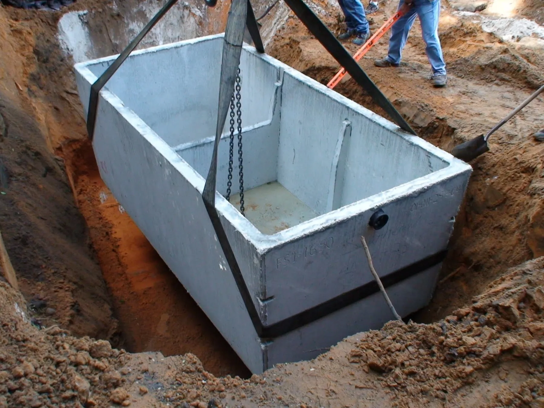 An open precast concrete septic tank hanging from nylon straps and chains as it is lowered into a freshly dug red clay pit.