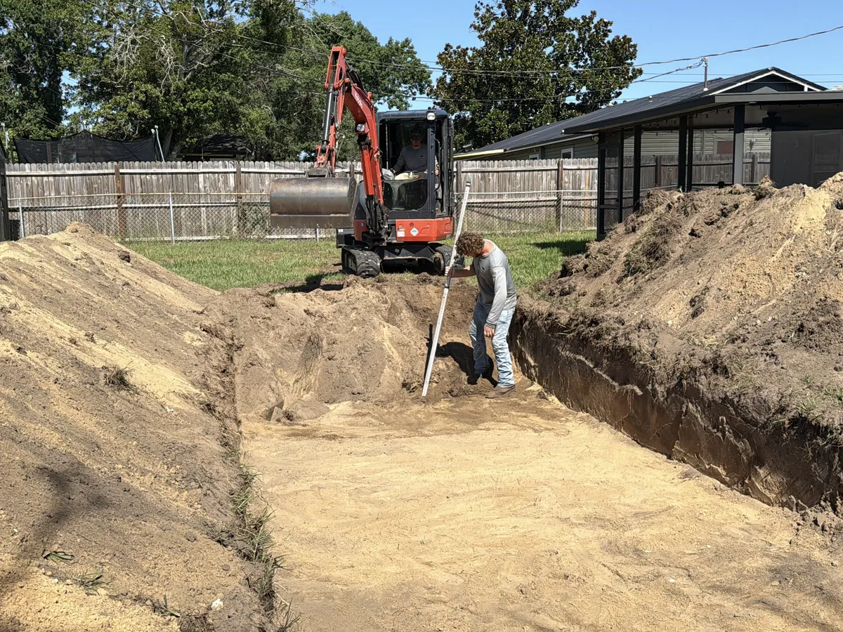A long drain field trench cut through sandy soil behind a house, with a mini excavator at the far end of the run.