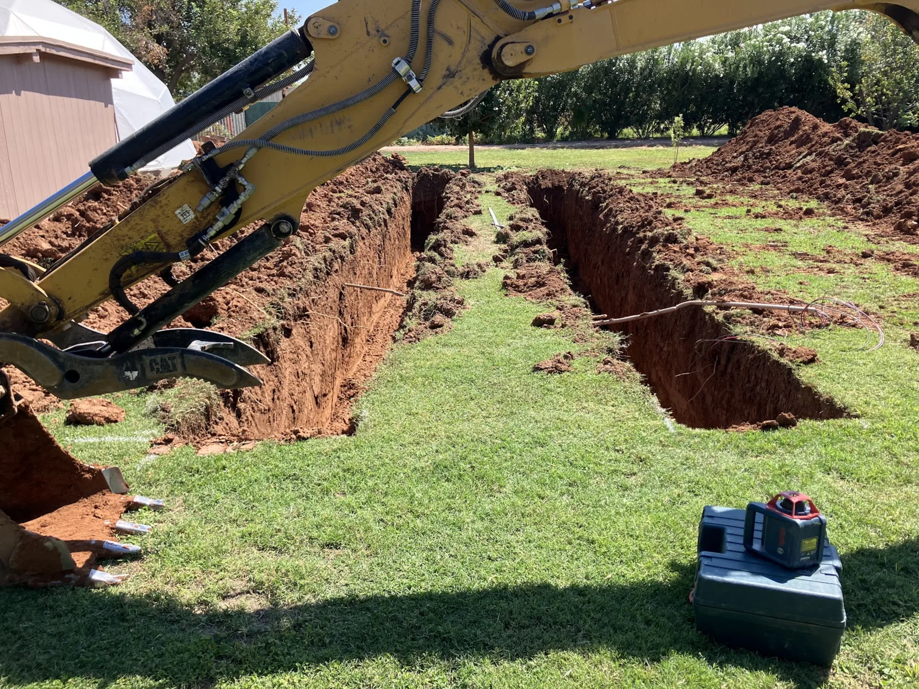 Two freshly cut drain field trenches in red clay running across a green lawn, with an excavator arm and a laser level box at the edge of the work.