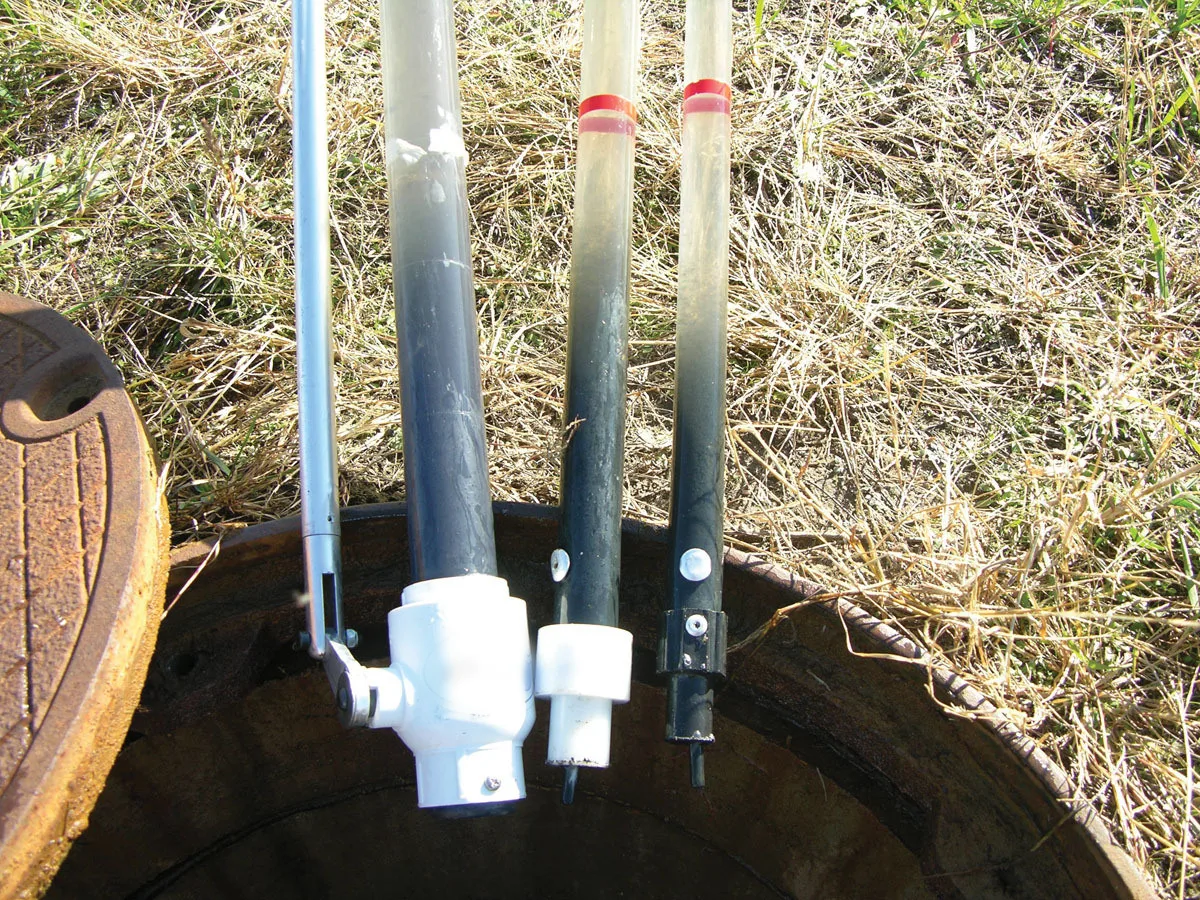 Three clear sludge sampling tubes resting across the open riser of a septic tank, each one holding a dark column of settled sludge